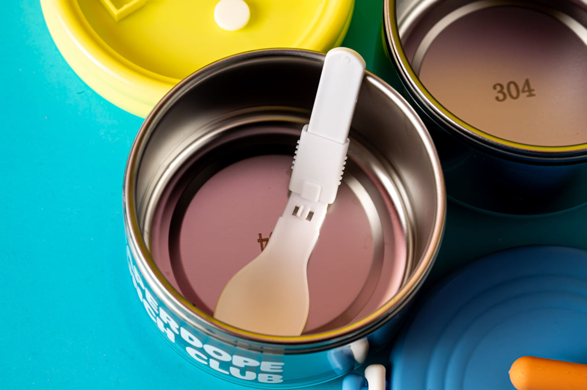 A close-up of a metal can with a translucent white plastic spoon inside, set on a bright turquoise background with other colorful lids and another can partially visible.